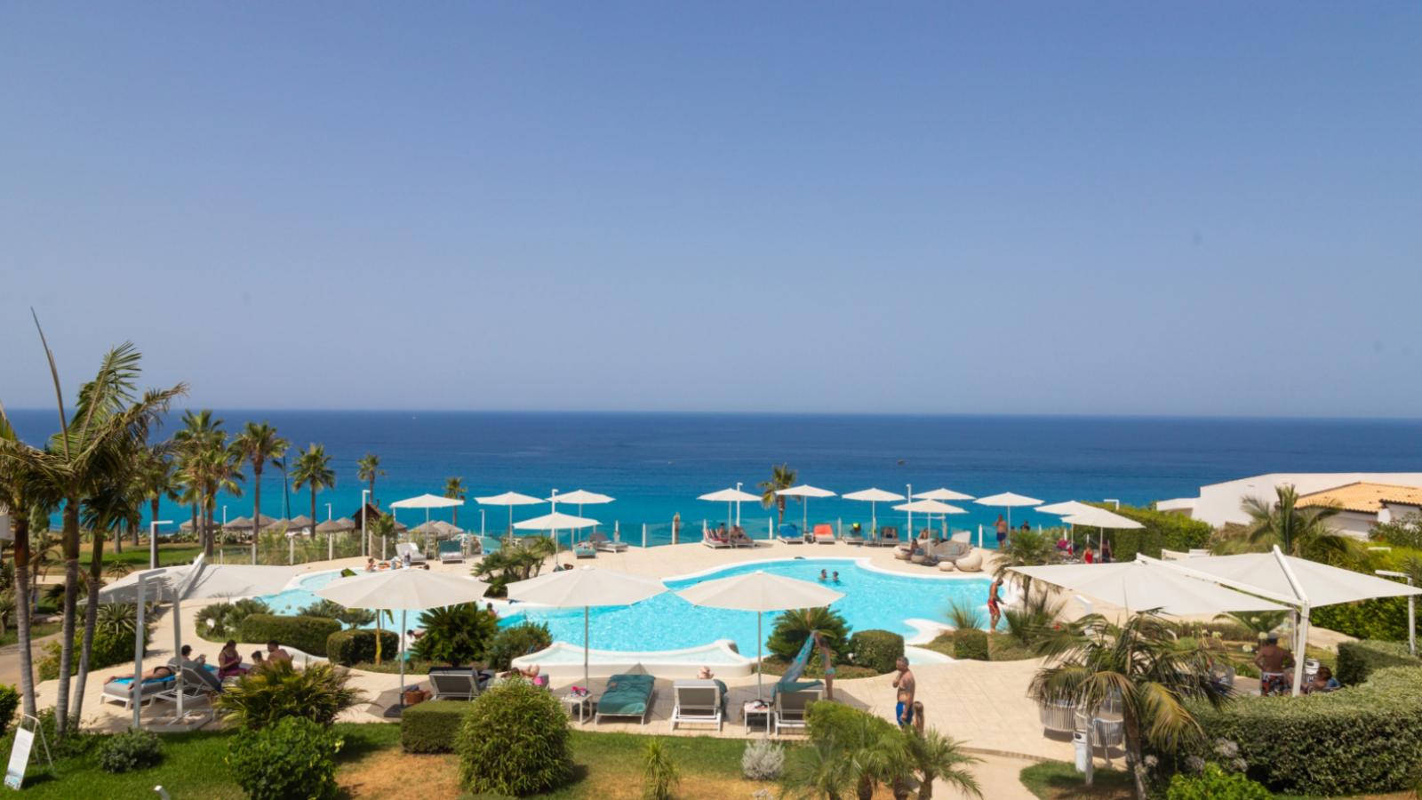 Resort con piscina e vista mare, palme e ombrelloni bianchi.