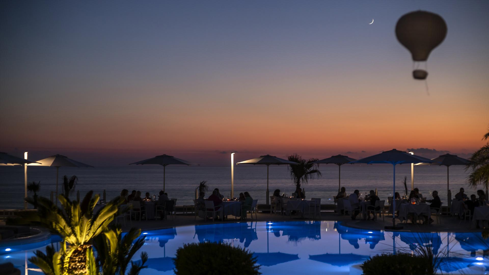Coucher de soleil sur la mer avec piscine illuminée et des gens dînant.