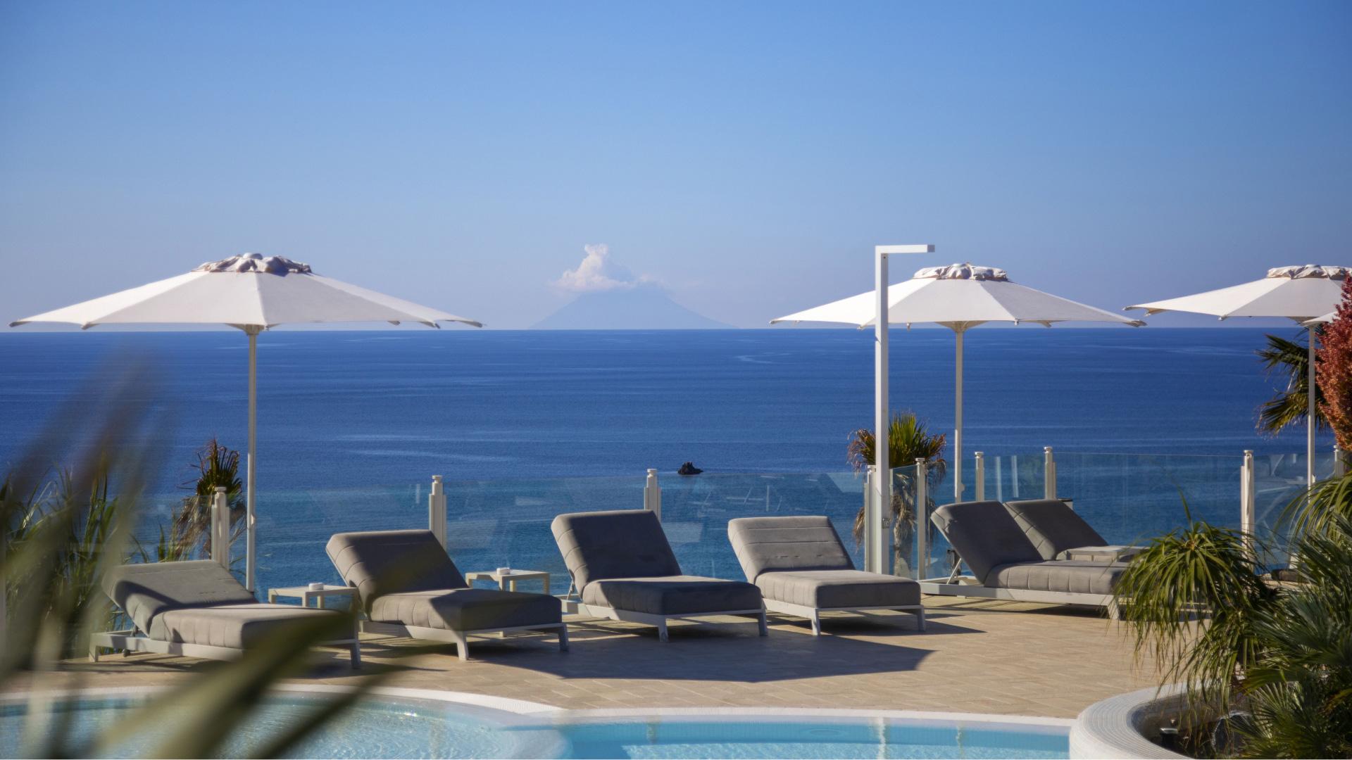 Pool with loungers and umbrellas, sea view and distant volcano.