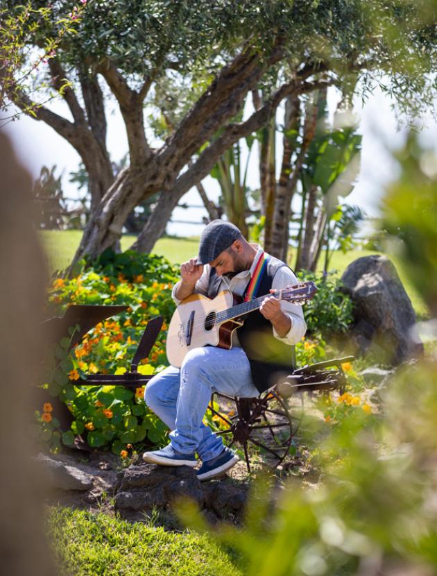 Uomo seduto suona la chitarra in un giardino fiorito.