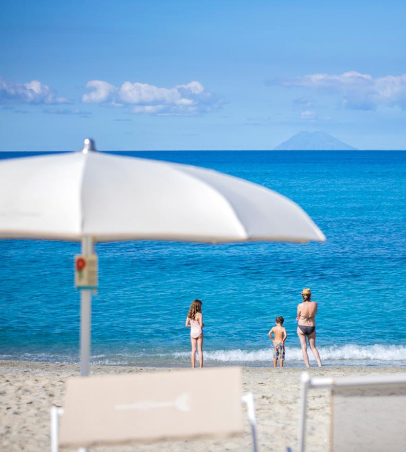 Spiaggia con ombrelloni, persone in mare e isola all'orizzonte.
